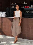 A woman stands in a cafe setting wearing a Nawa White ribbed knit tank top and an asymmetrical taupe-brown pleated midi skirt.