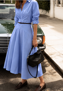 A woman wearing a light blue midi shirt dress with a thin dark brown belt, holding a black leather shoulder bag.